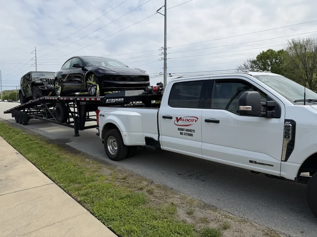 Velocity Logistics branded pickup towing an open car hauler loaded with a Tesla and a Jeep Wrangler