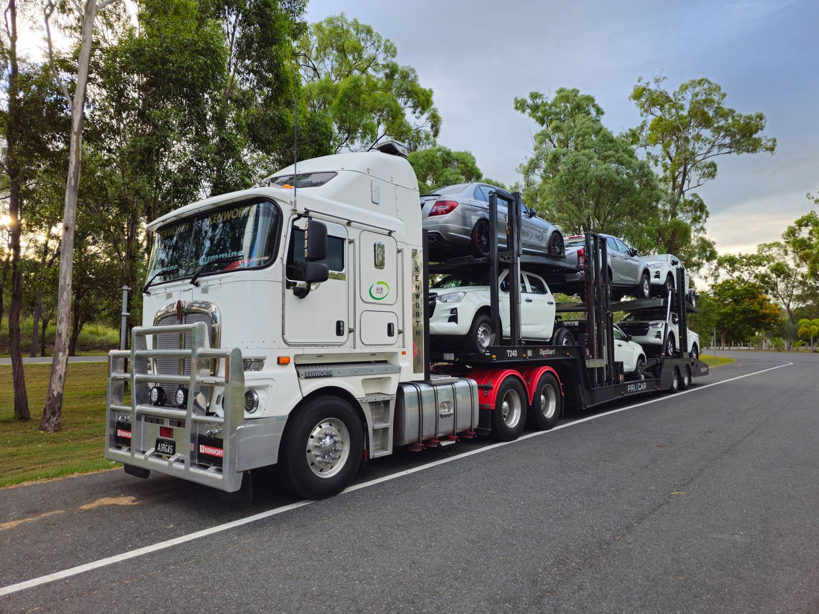 A multi-car open transport carrier loaded with vehicles driving on a tree-lined road