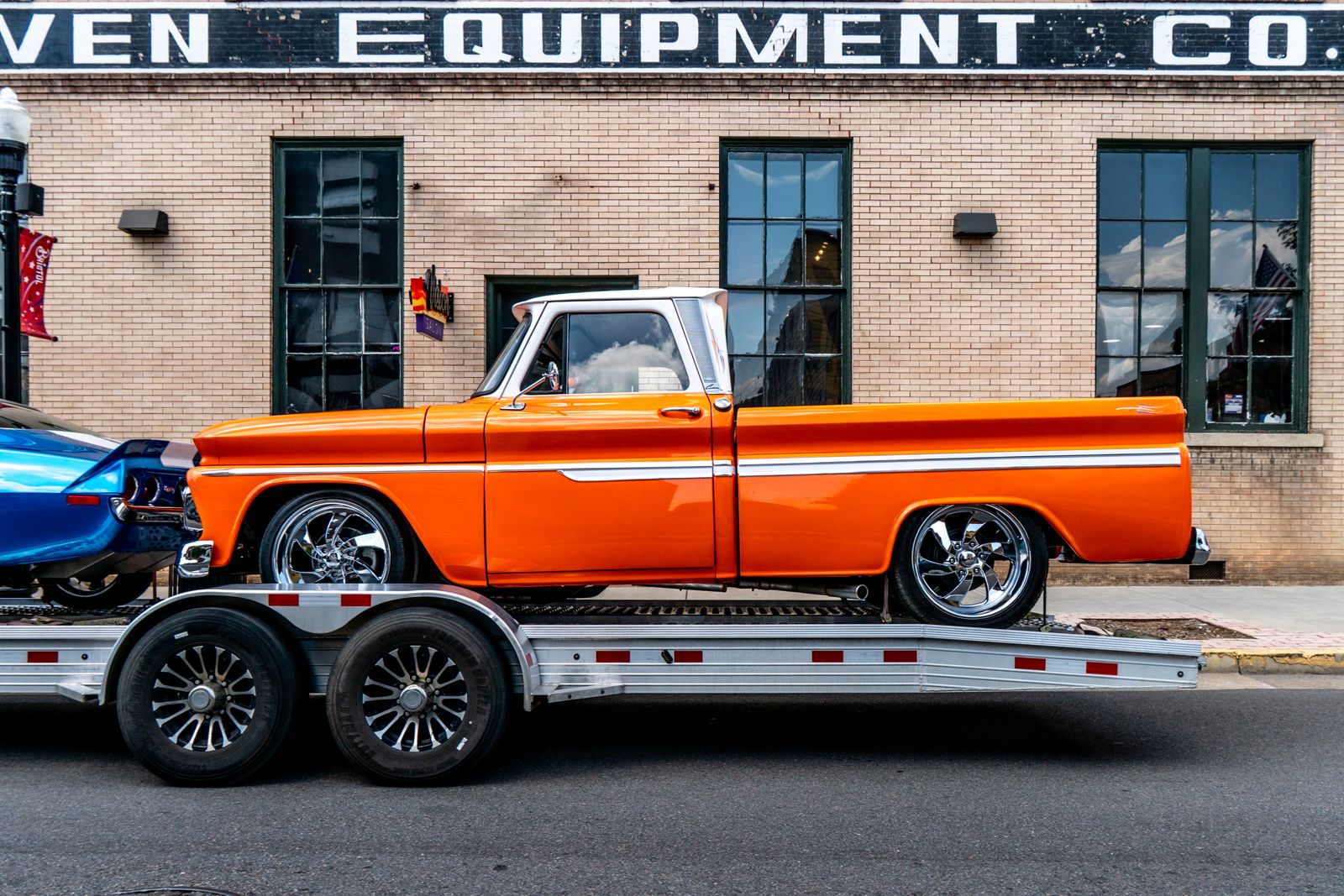 A restored classic orange pickup truck secured on an open car trailer in a downtown setting