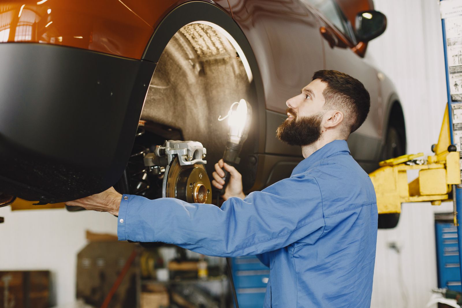 A technician using a flashlight to inspect a vehicle's brakes and wheel on a lift